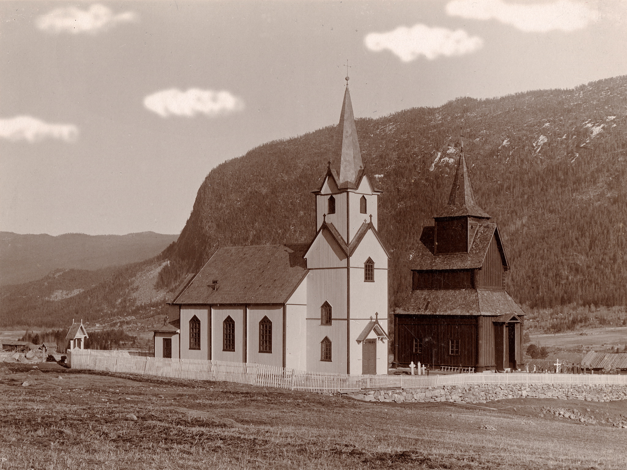 Da Torpo nye kyrkje (til venstre) stod ferdig i 1880 ville dei rive stavkyrkja (til høgre) frå slutten av 1100-talet. Dei starta med å rive koret men da Fortidsminneforeningen kjøpte kyrkja blei rivinga stansa og skipet bevart. Skyene er teikna inn på biletet noko som er typisk for mange av bileta til C. Christensen Thomhav (1857 - 1937).Foto: C. Christensen Thomhav / Riksantikvaren