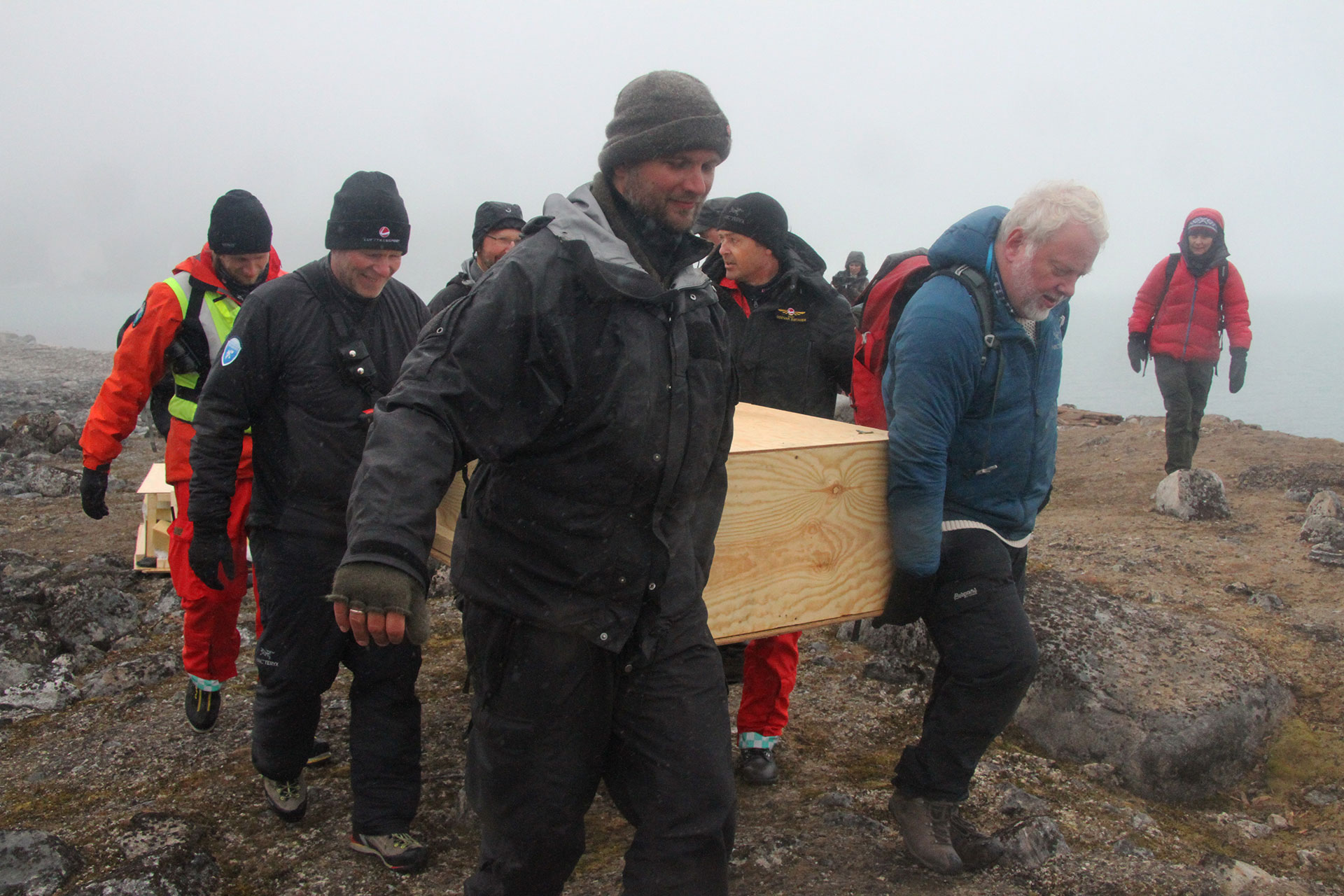 Bilde hvor kisten som inneholder restene fra en av hvalfangergravene bæres til helikopteret som skal frakte den til Svalbard museum. Inne i den nye kisten ligger hele den gamle graven, inkludert restene av gammel kiste og omkringliggende jord. Foto: Siri Wolland, Riksantikvaren