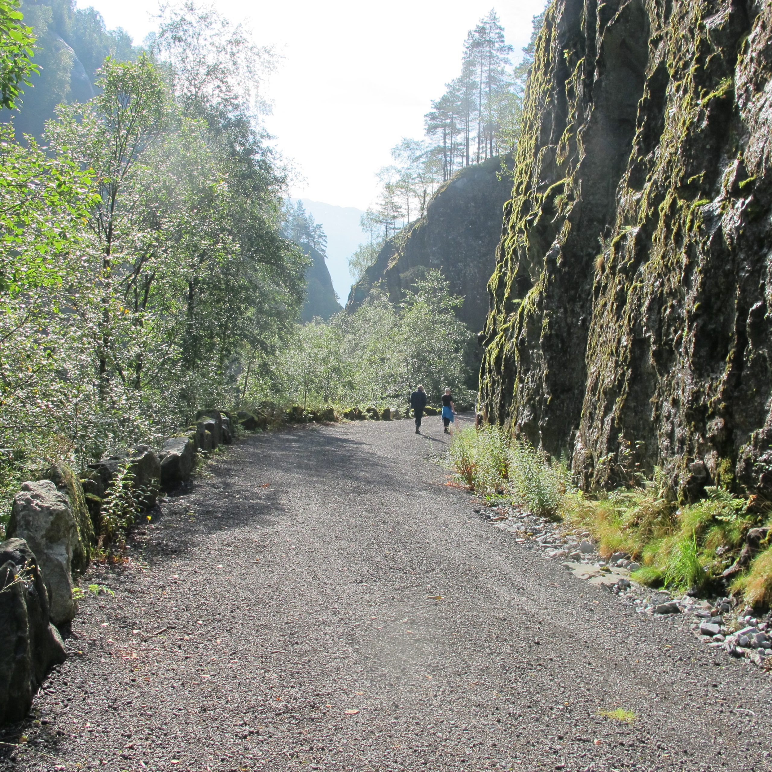 Bildet viser en gruslagt vei inntil fjellside og med steinbelagt kant på venstre side. Den er omkranset med grønne trær og vegetasjon, i enden av veien ser man to turgåere.