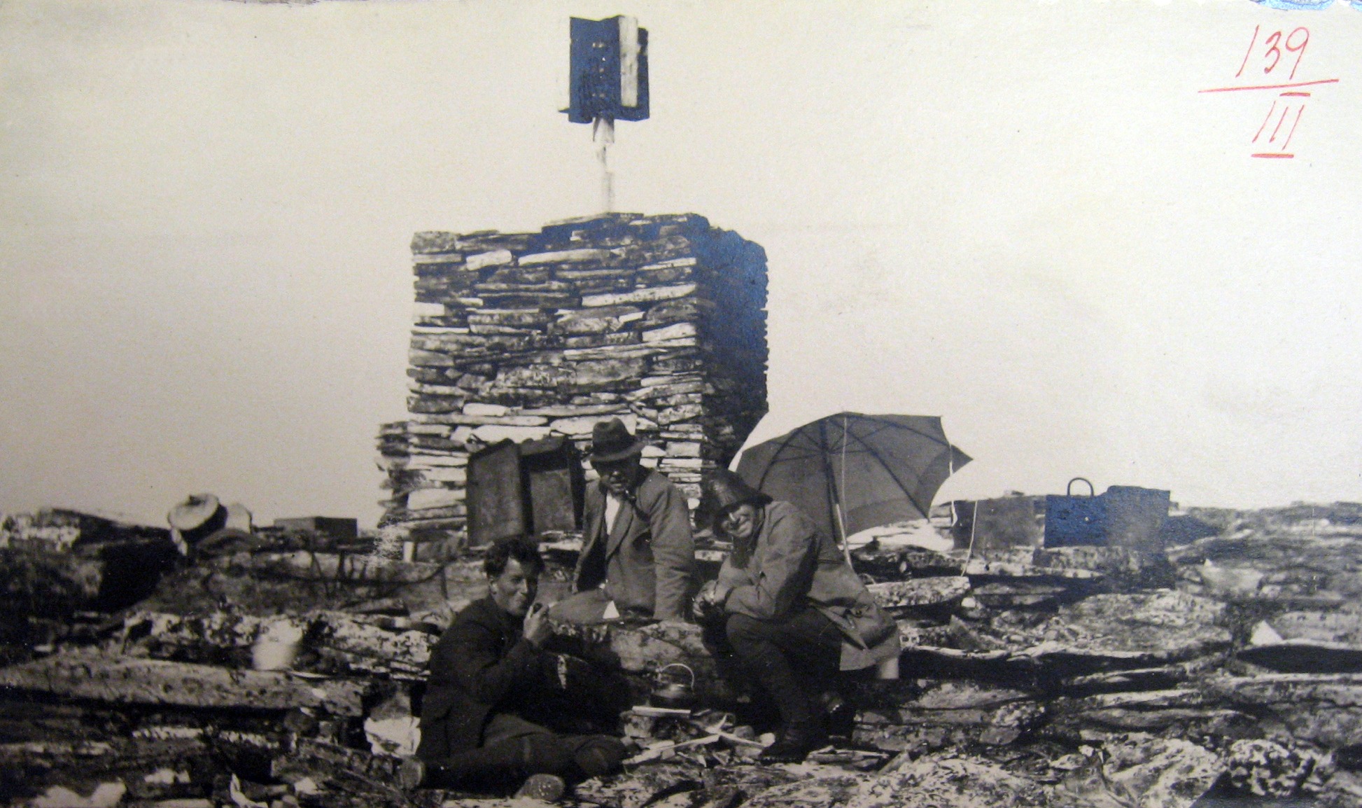 Bildet ser gammelt ut, og viser tre menn sittende foran med diverse utstyr plassert rundt steinvarden.