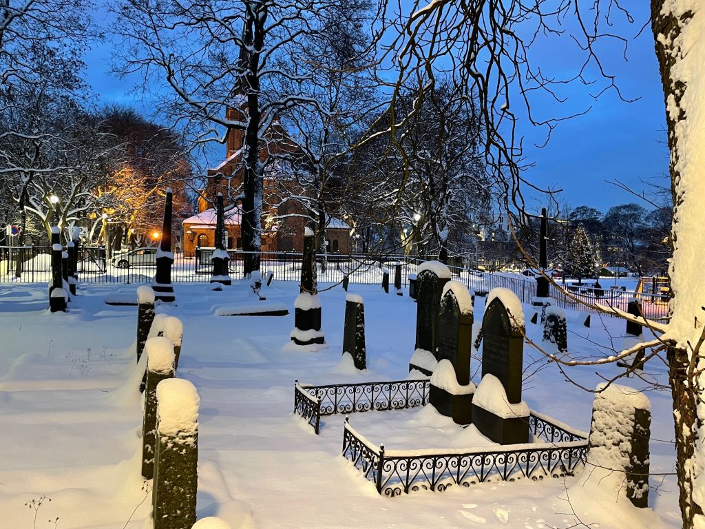 Bildet viser den jødiske gravlunden med gravstøtter, og med Sofienberg kirke i bakgrunnen. Bildet er tatt i grålysningen i vinterlandskap.