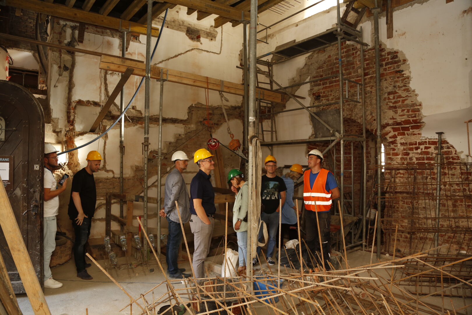 En gruppe mennesker med beskyttelsesutstyr på befaring i innsiden av et bygg under restaurering, med stilaser og utstyr.