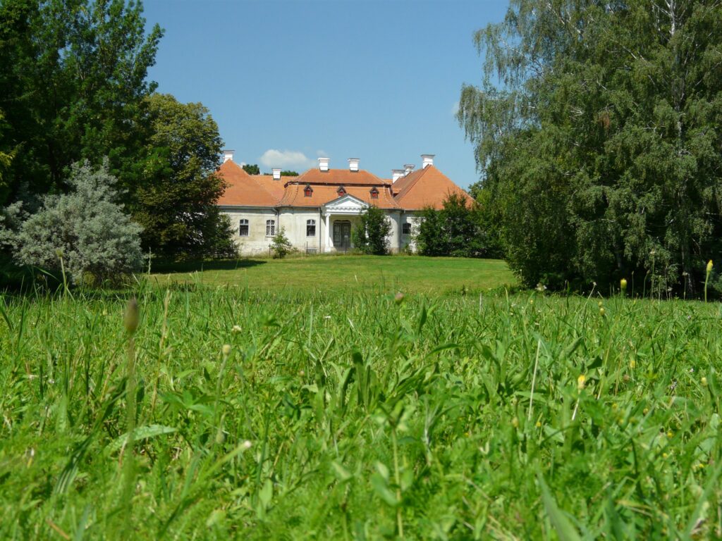 En herregård i gul murstein med rødt tak, beliggende på en stor grønn plen med grønne trær rundt.