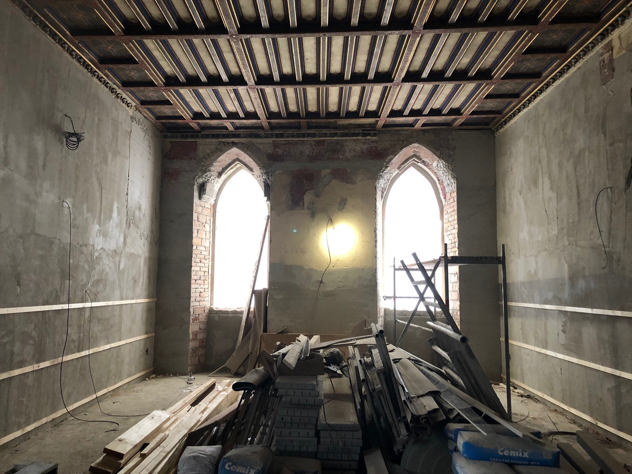 Innsiden av et rom under renovering, med stilaser, planker og murstein i stabler. Et eksponert tretak og høye, gotiske vinduer uten glass.