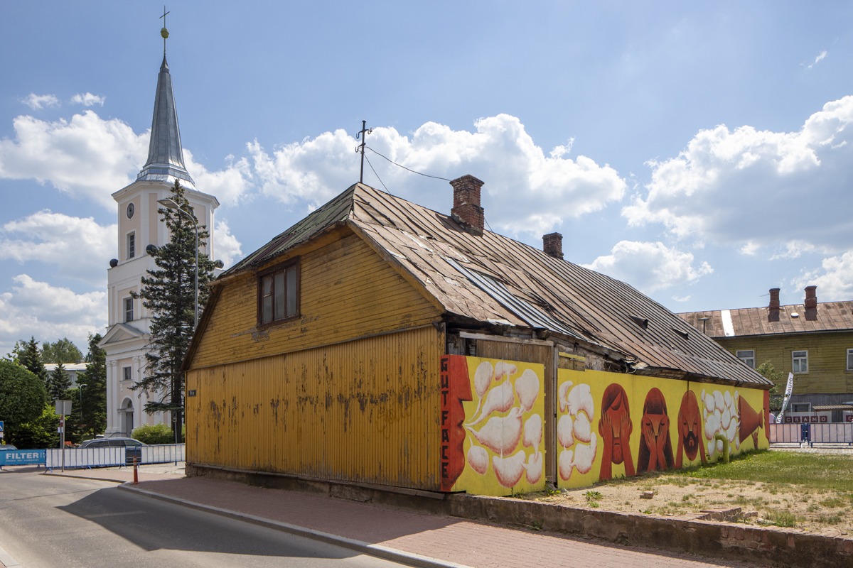 En kirke i bakgrunnen, i forgrunnen et gammelt, skjevt gult trehus med malte sponplater foran den ene langveggen.