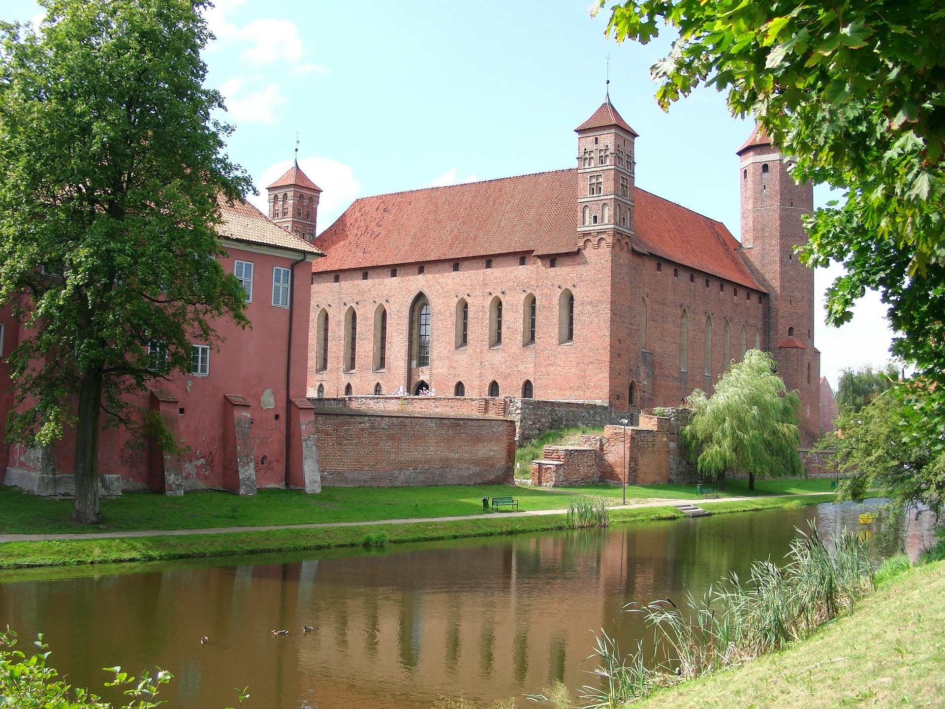 Et stort slott i rød murstein, med fire tårn og spir, beliggende ved en elv med grønne plener og trær foran.