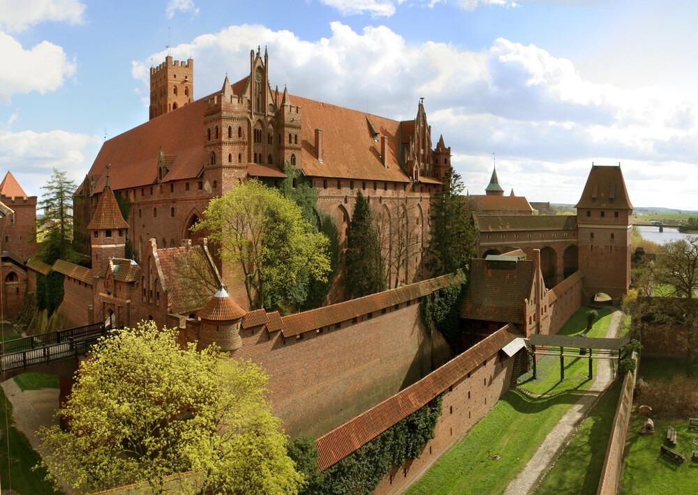 Et gigantisk slottskompleks i rødbrun murstein, med flere yttergående murvegger, tårn og borggård. Omgitt av grønne plener, stier og en elv i bakgrunnen.