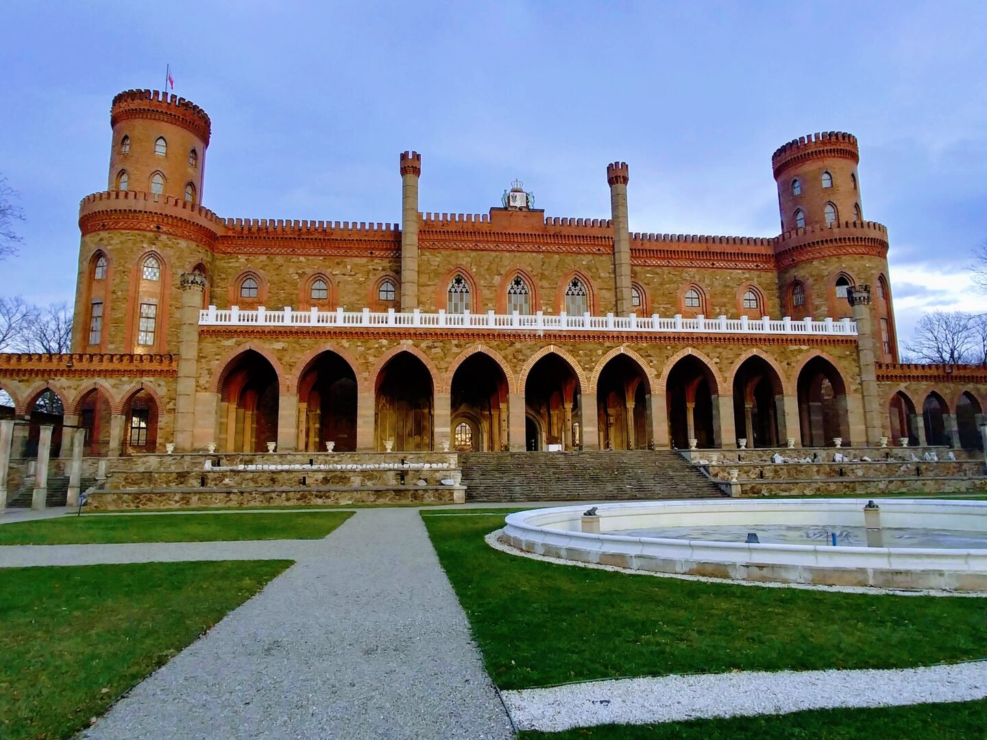 Et palass i gotisk stil med to tårn, bygget i rødbrun murstein med grønn plen og fontene i front.