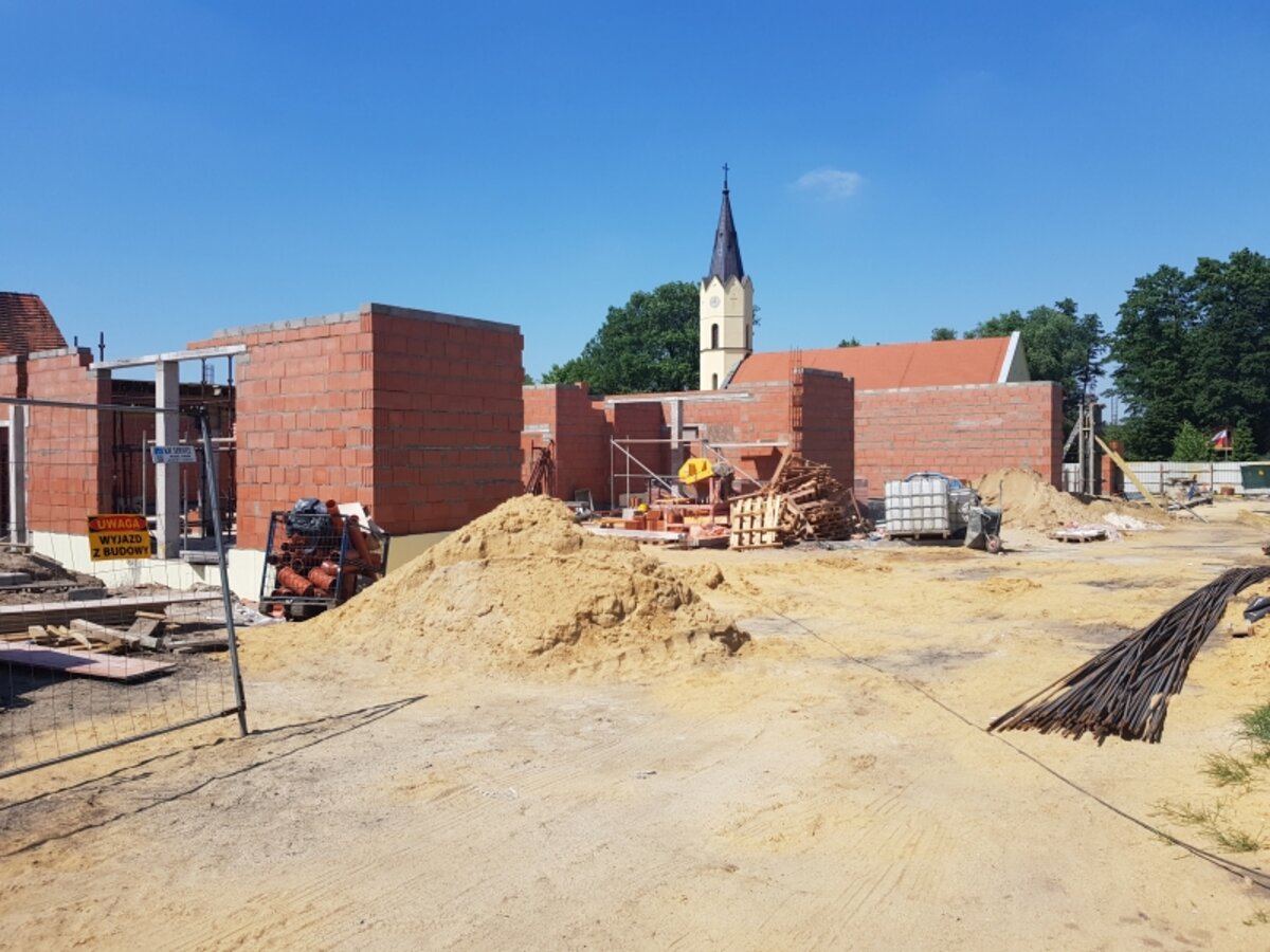 En byggeplass med materialer og maskiner, delvis konstruerte vegger i rød murstein, en kirke i bakgrunnen.