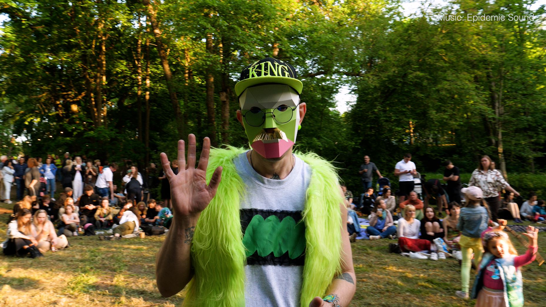 En mann med fargerik maske og grønn fuskepelsvest, meg sort og grønn caps hvor det står "King", som vinker til kameraet. Andre publlikummere i bakgrunnen som sitter eller står på plenen.