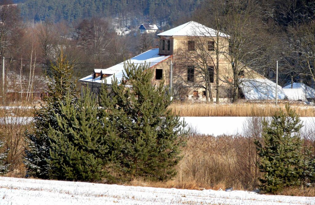 En bygning i tre etasjer med to sidefløyer, stående på et jorde i vinterlandskaå med snø på bakken og grantrær i forgrunnen.