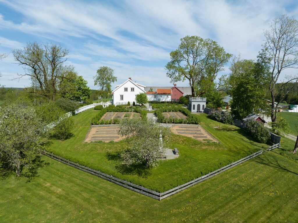 På bildet ser man hagen på Spydeberg prestegård i Østfold, med det hvite trehuset i bakgrunnen. Hagen har flere arealer for dyriking og er satt i stand og godt ivaretatt.