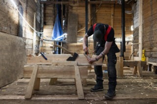 Bildet viser en mann bøyd over en høvelbenk, der han utfører arbeide med tradisjonelle verktøy på en større planke